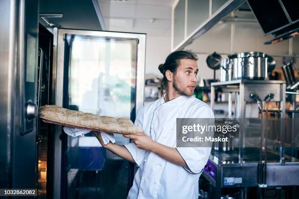 male chef putting bread in oven while looking away at commercial kitchen - baking bread stock pictures, royalty-free photos & images