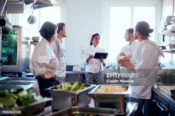 mid adult female chef holding digital tablet while discussing with team in kitchen - food and drink industry stock pictures, royalty-free photos & images
