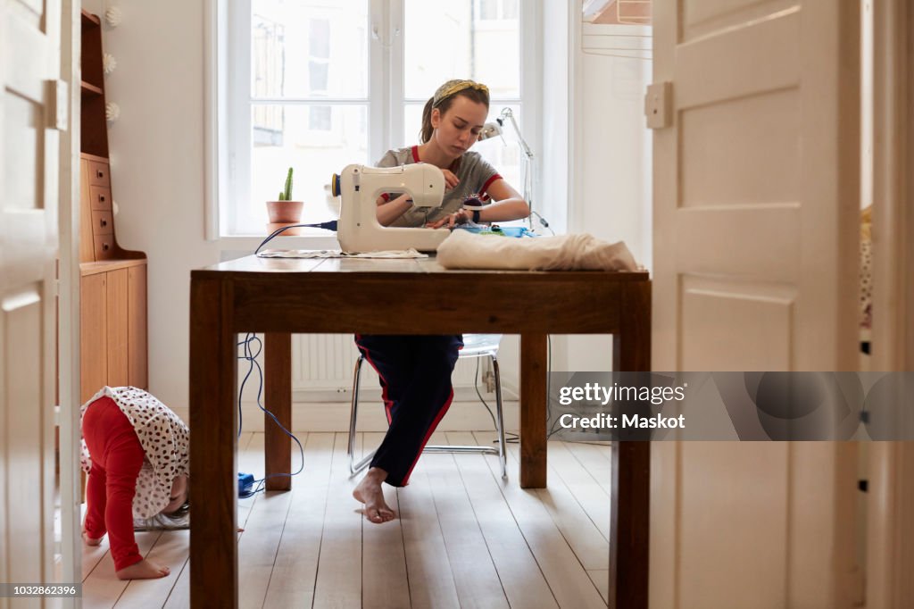 Tailor sewing at table while daughter playing on floor seen through doorway at home