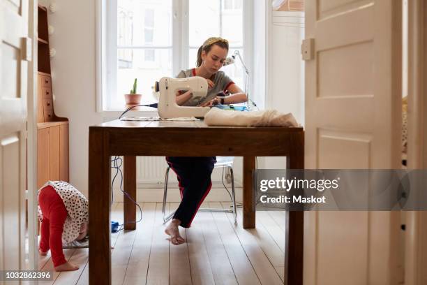 tailor sewing at table while daughter playing on floor seen through doorway at home - sewing machine stock pictures, royalty-free photos & images