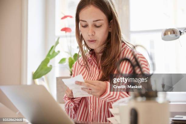 young woman examining financial bill while using laptop at home - accesorio financiero fotografías e imágenes de stock