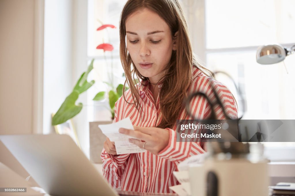 Young woman examining financial bill while using laptop at home