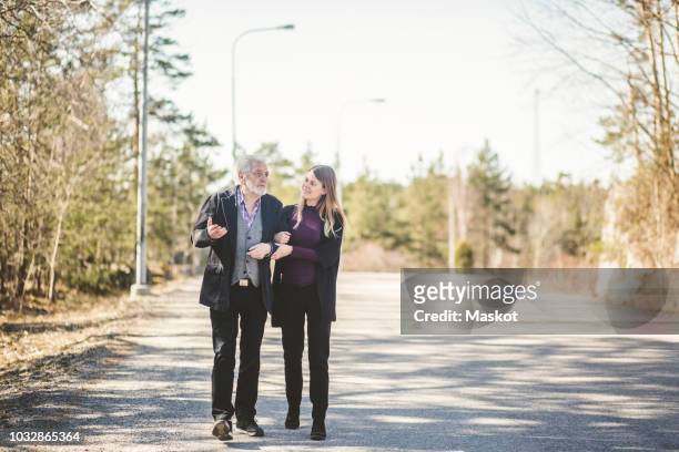 full length of young woman walking arm in arm with grandfather on road - old man young woman stock pictures, royalty-free photos & images