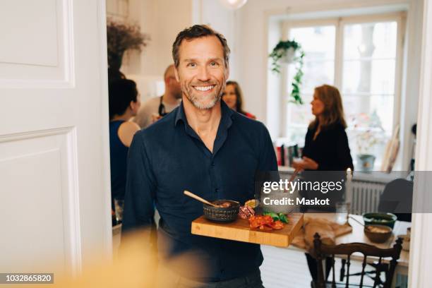 portrait of smiling mature man holding food in serving tray while standing against friends at party - dinner party at home stockfoto's en -beelden