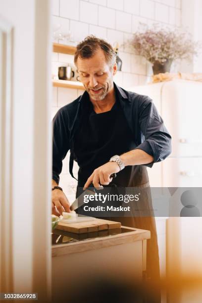 mature man cutting cheese on board at kitchen counter - man cooking photos et images de collection