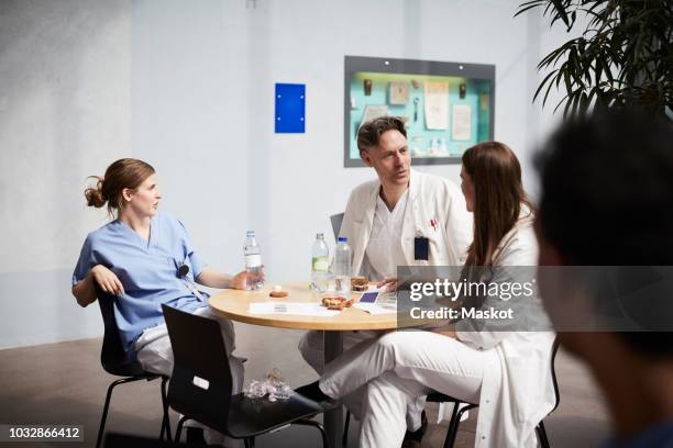 confident mature doctor discussing with female coworkers while sitting at table in hospital cafeteria - three female doctors stock pictures, royalty-free photos & images