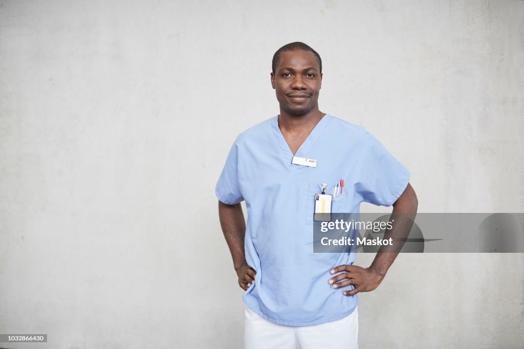 Portrait of confident mid adult male nurse standing with hands on hips in corridor at hospital