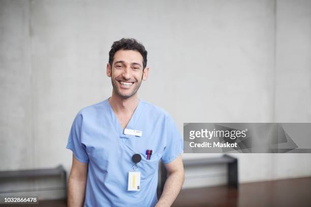 portrait of smiling young male nurse in blue scrubs standing against wall at hospital - male medical professional stock pictures, royalty-free photos & images