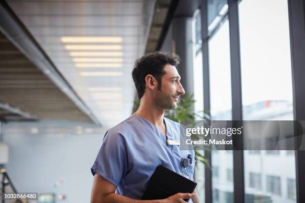 thoughtful young male nurse holding digital tablet while looking through window at hospital - male medical professional stock pictures, royalty-free photos & images