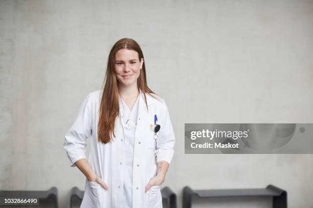 portrait of smiling young female brunette doctor standing with hands in pockets at hospital - laborkittel stock-fotos und bilder