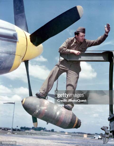 Cpl. Lloyd Shumway, standing atop a 500 lb. Bomb, directs a crane into placing the bomb on the wing of a North American P-51, at an unidentified air...