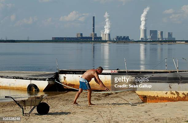 Caretaker sweeps debris from a beach next to a small mooring pier for boats at Baerwalder See lake as exhaust rises from the Boxberg coal-burning...