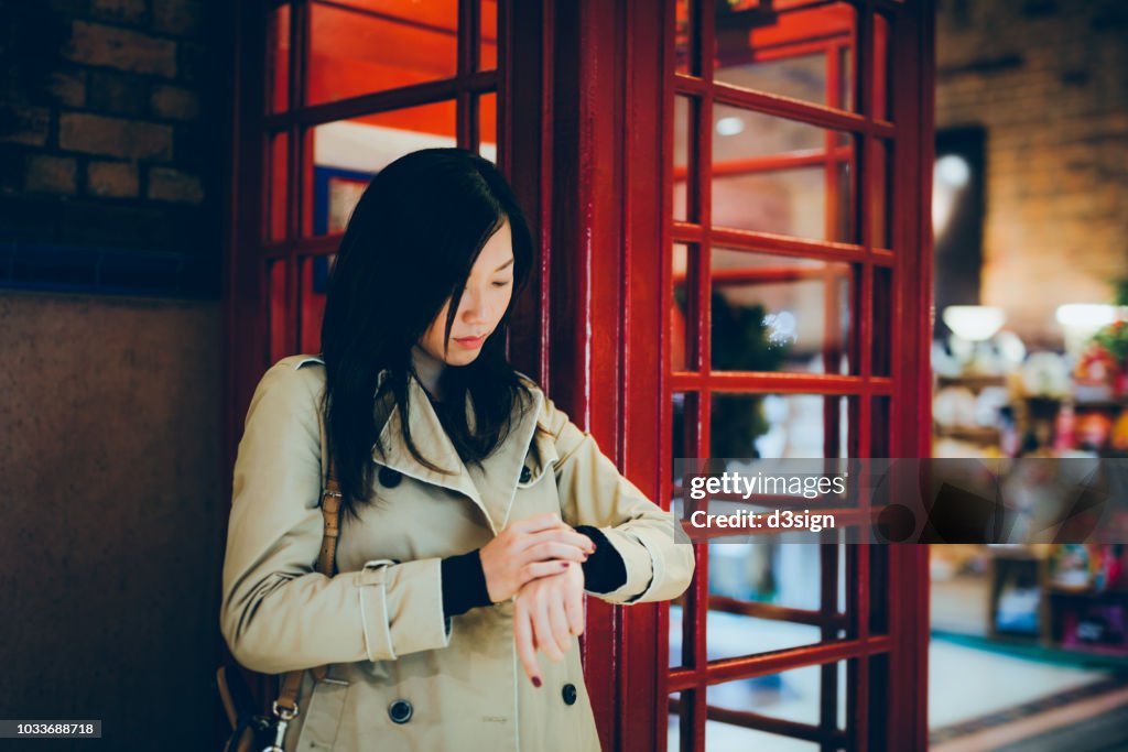 Young woman checking time and waiting next to red telephone booth in city