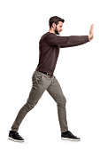 A bearded man in casual clothes tries to push a heavy object with both arms with one leg put in front for balance.