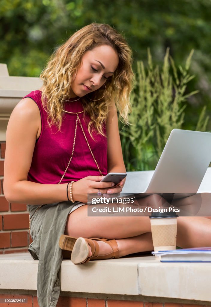A beautiful young female university student sits working on her laptop outside on the campus, and texting on her smart phone