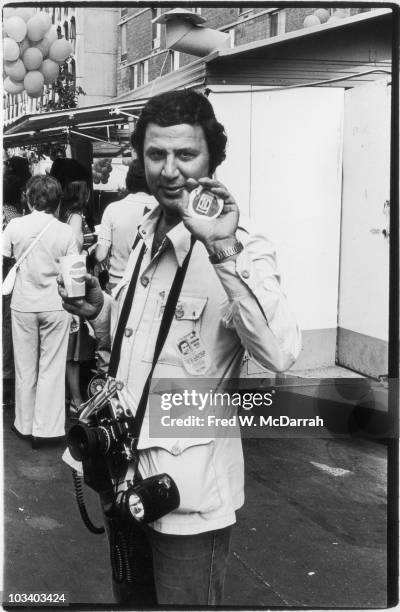 Portrait of American photogragher Ron Galella as he poses on the street and holds up a printed picture of an apple that sports a Pepsi logo; he holds...