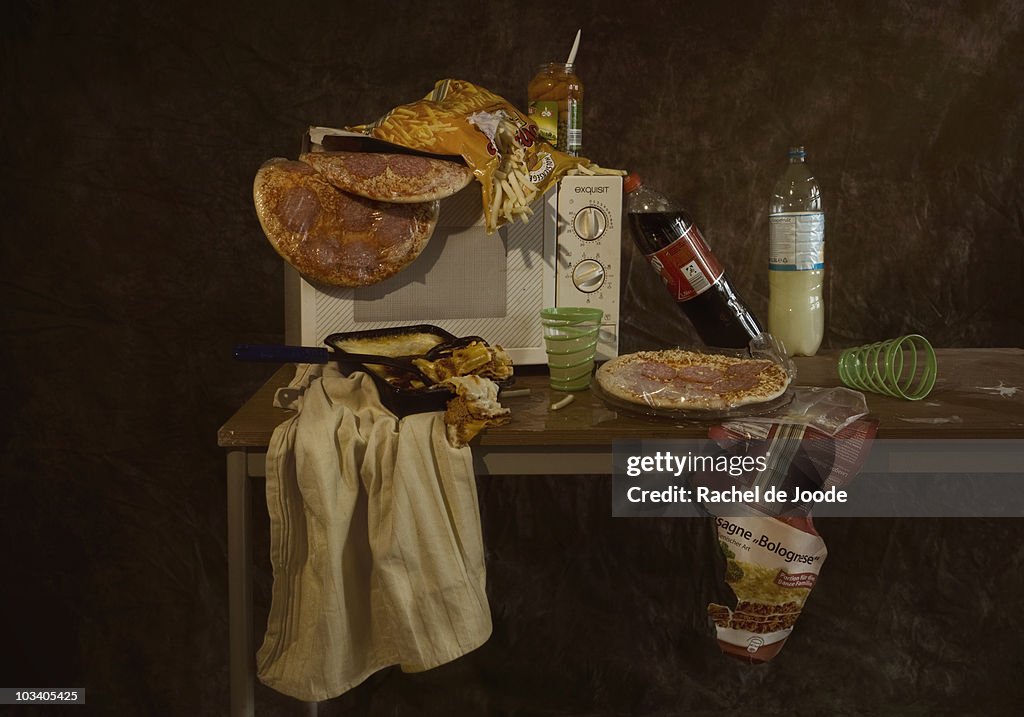 A table with a microwave and various frozen pizzas and frozen lasagna, still life