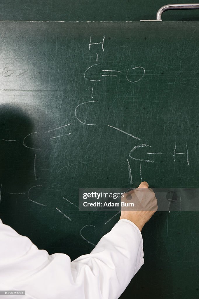 A chemistry teacher writing a formula on a chalkboard, focus on hand