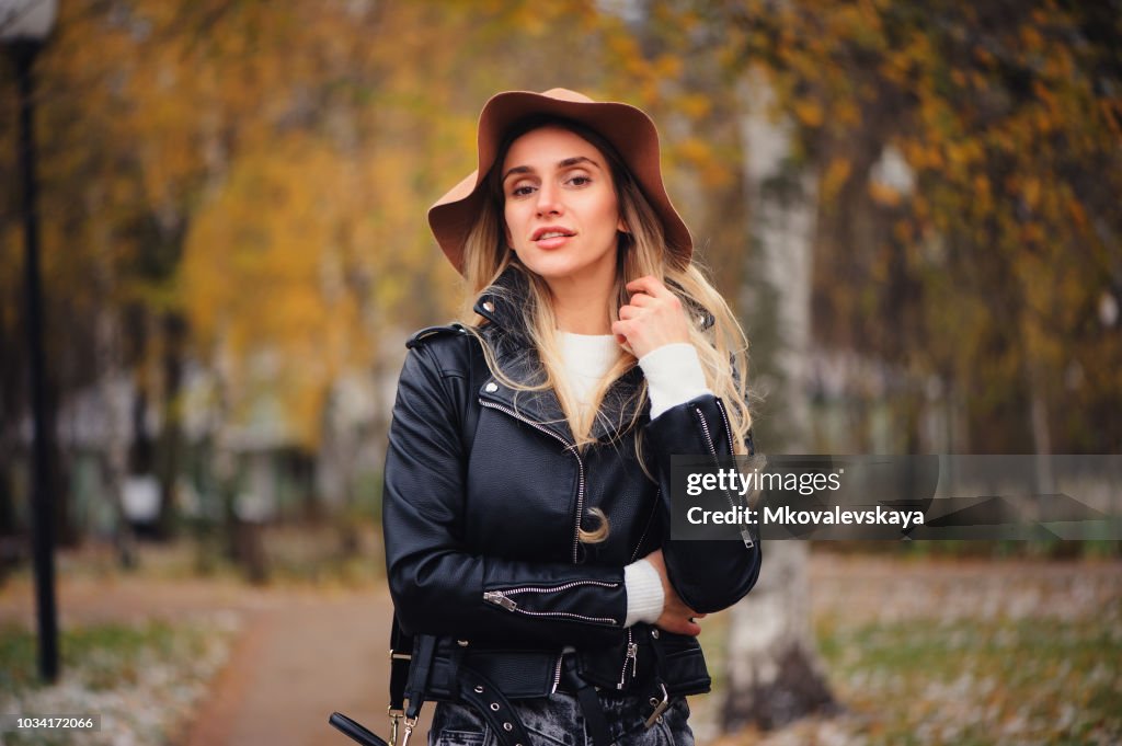 Fashion autumn portrait of young happy woman walking outdoor in fall park in hat and leather jacket