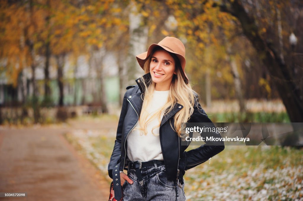 Fashion autumn portrait of young happy woman walking outdoor in fall park in hat and leather jacket
