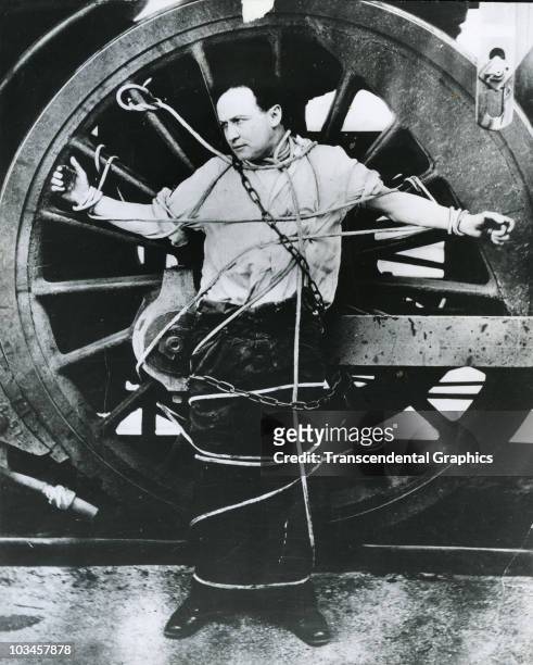 In a publicity stunt, magician and escape artist Harry Houdini chains himself to a locomotive wheel for an escape attempt, ca.1910.