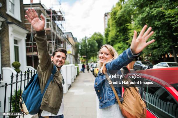 a young couple with backpacks walking up the street, looking back nad waving at somebody. - female waving on street stock pictures, royalty-free photos & images