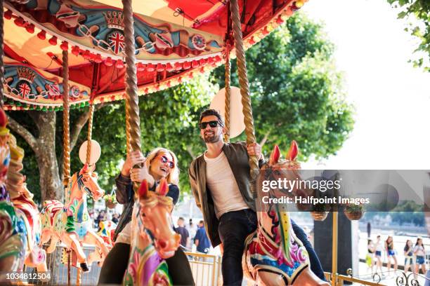 a happy young couple with sunglasses riding on carousel horses. - draaimolen stockfoto's en -beelden