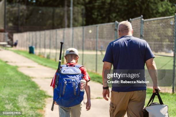 father and son walking on paved path to baseball diamond carrying baseball equipment - carrying sports bag foto e immagini stock
