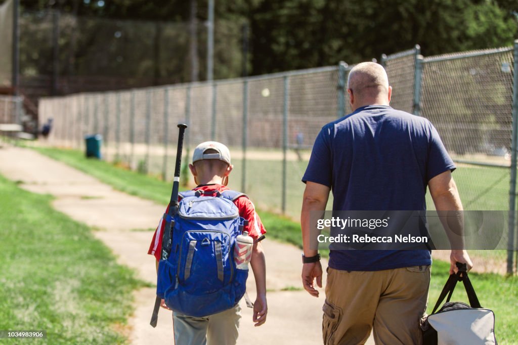 Father and son walking on paved path to baseball diamond carrying baseball equipment