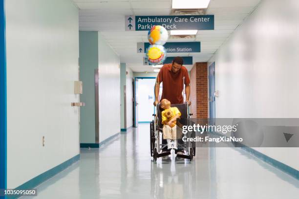 padre africano empujando a hijo en silla de ruedas por el pasillo del hospital - tienda de regalos fotografías e imágenes de stock