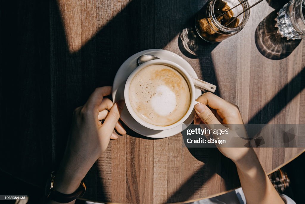 Top view of woman sitting by the window in coffee shop enjoying the warmth of sunlight and drinking coffee