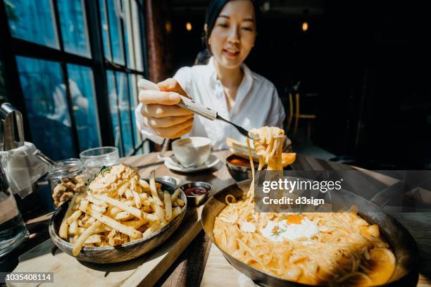 young asian woman sitting at a table by the window enjoying the warmth of sunlight and having meal joyfully in a restaurant - asian couple dining stockfoto's en -beelden