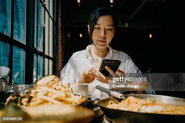 young asian woman checking smartphone while having meal in a restaurant - dinner program stock pictures, royalty-free photos & images