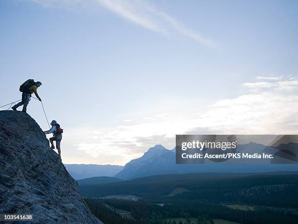 man belays woman to top of cliff above mtn valley - zekeren stockfoto's en -beelden