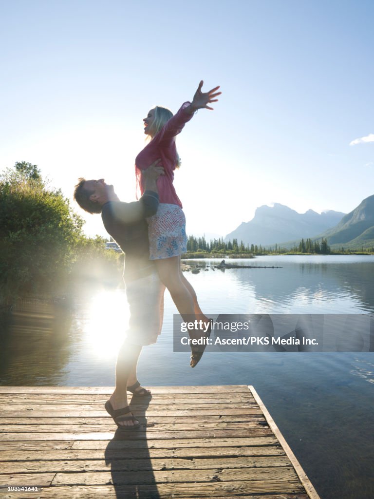 Man lifts woman into air on wooden wharf, mtns