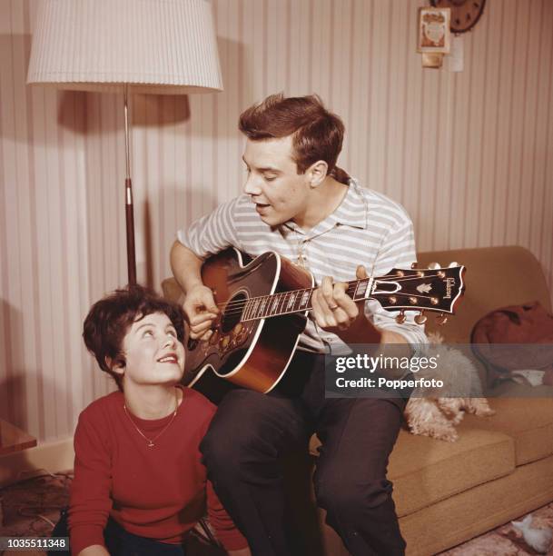 English pop singer Marty Wilde pictured playing a Gibson acoustic guitar with his wife Joyce in a living room at home in England in 1960.