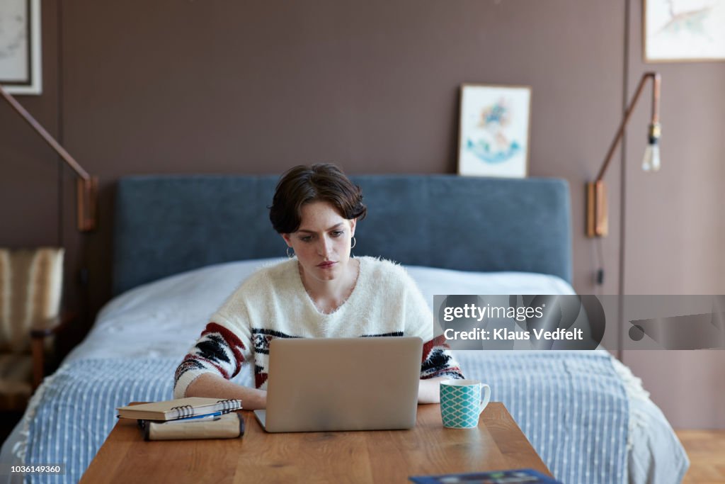 Cool young woman sitting in apartment working on laptop