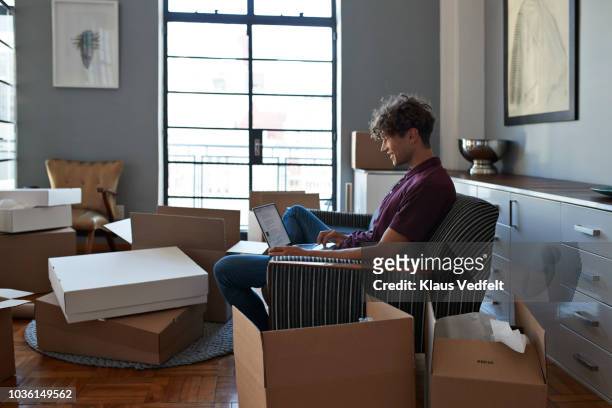 young man typing on laptop in apartment full of boxes - man with moving boxes authentic stockfoto's en -beelden