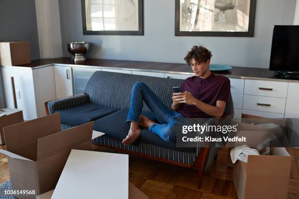 young man scrolling on smartphone in apartment full of boxes - classic mens day 1 stock pictures, royalty-free photos & images