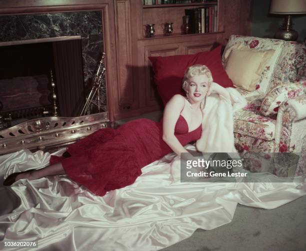 American actress Marilyn Monroe in a red brocade evening gown, reclining on white satin, with her head resting on a white fur coat, 1955.