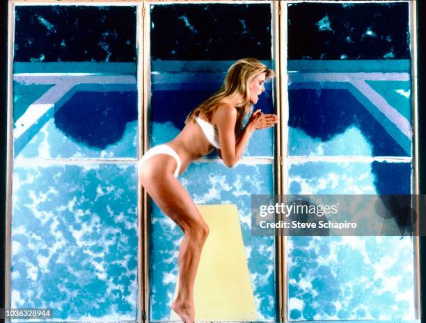 Portrait of American model Christie Brinkley, dressed in a white bikini, mines a dive as she poses in front of a David Hockney's 'Day Pool With Three...