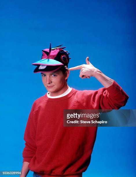 Portrait of Canadian actor Michael J Fox, in a red sweatshirt, as he points to a novelty baseball cap on his head, Los Angeles, California, 1983.