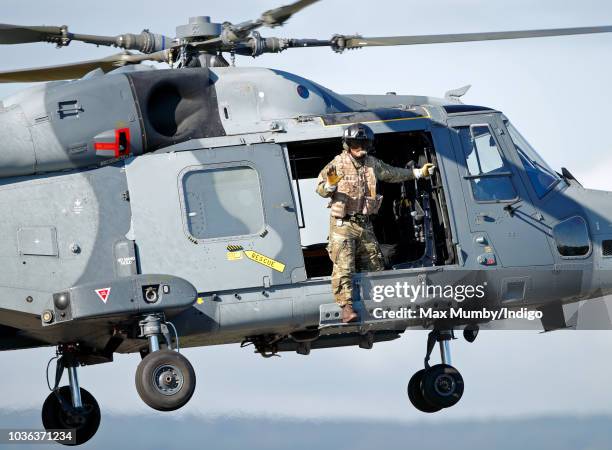 Royal Marines Commando hangs out of the open door of a Royal Navy Wildcat Maritime Attack Helicopter as it departs The Royal Marines Commando...