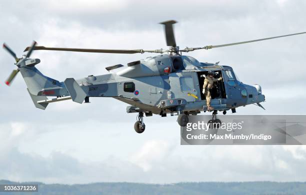 Royal Marines Commando hangs out of the open door of a Royal Navy Wildcat Maritime Attack Helicopter as it departs The Royal Marines Commando...