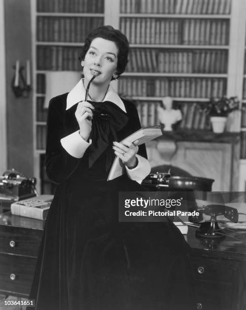 Actress Rosalind Russell pictured seated on the edge of a desk while putting the arm of a pair of glasses to her lips with one hand and holding a...