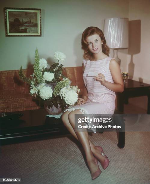 Actress Ann-Margret pictured sitting on a coffee table, wearing a pink and white dress, alongside a vase of flowers, USA, circa 1965.