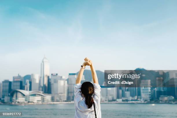 rear view of woman traveller enjoying her time in hong kong, taking a deep breath with hands raised against victoria harbour and city skyline - travel market asia stock-fotos und bilder