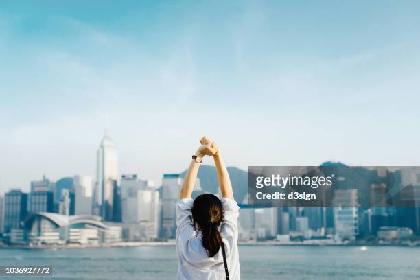 rear view of woman traveller enjoying her time in hong kong, taking a deep breath with hands raised against victoria harbour and city skyline - prosperity stock pictures, royalty-free photos & images
