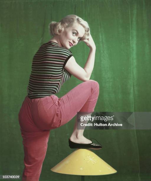 Actress Shirley Eaton pictured with her foot resting on a lampshade, wearing red trousers and a striped top, circa 1955.