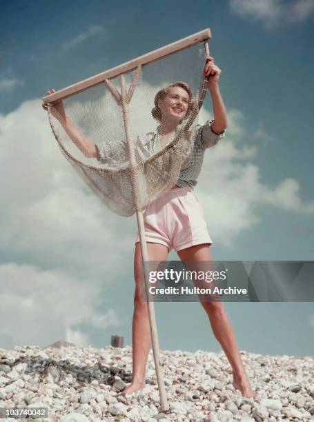 Model wearing white shorts and green gingham shirt poses with a fishing net on a pebble beach, Great Britain, circa 1955.